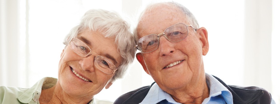 Close up photo of an older white couple with their heads together smiling and looking at the camera
